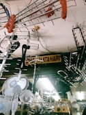 Numerous metallic antennas and fans are hanging from the ceiling of a shop. The items appear to be stacked closely together, creating a cluttered visual effect. There is a signboard partially visible in the background.