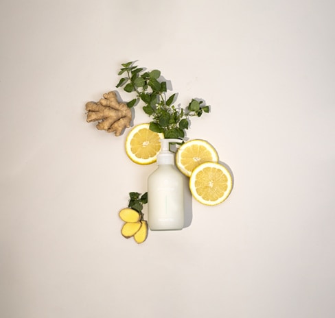 A neatly arranged composition featuring slices of lemon, fresh green leaves, a piece of ginger root, and a white bottle, all placed against a light background. The layout creates a refreshing and natural aesthetic.