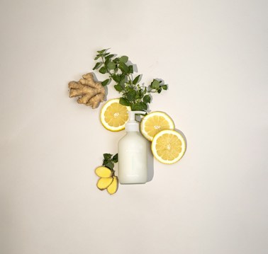 A neatly arranged composition featuring slices of lemon, fresh green leaves, a piece of ginger root, and a white bottle, all placed against a light background. The layout creates a refreshing and natural aesthetic.