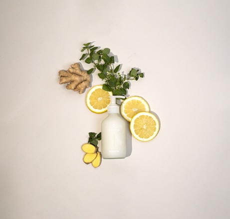 A neatly arranged composition featuring slices of lemon, fresh green leaves, a piece of ginger root, and a white bottle, all placed against a light background. The layout creates a refreshing and natural aesthetic.
