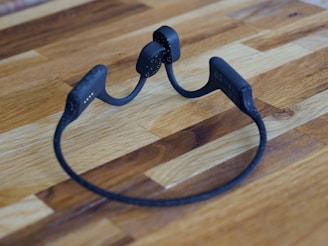 A pair of black wireless headphones with a flexible neckband are placed on a wooden surface. The headphones have a sleek and modern design.