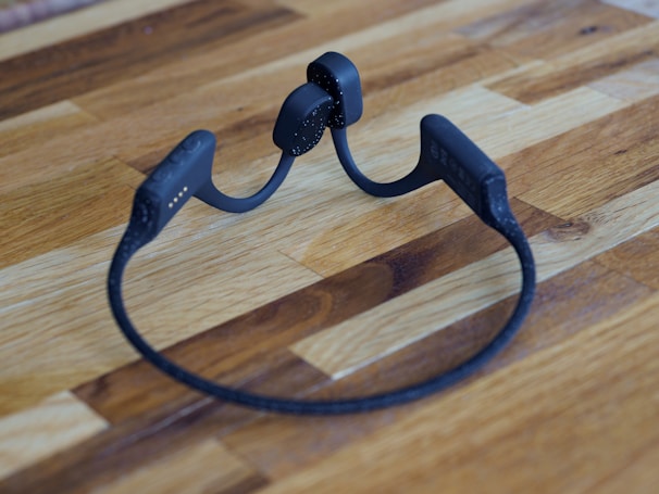 A pair of black wireless headphones with a flexible neckband are placed on a wooden surface. The headphones have a sleek and modern design.