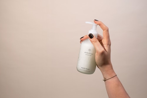 A hand with dark nail polish holds a white bottle labeled 'everdrop natural care' with a pump dispenser. The background is plain and neutral-colored.