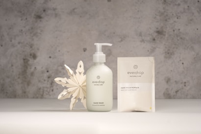 A minimalist product display featuring a hand wash bottle and a hand wash powder sachet from the brand Everdrop. The items are placed on a light grey surface with a textured, grey concrete background. Beside the products is a decorative white paper snowflake, adding an artistic touch to the scene.