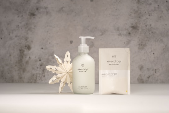 A minimalist product display featuring a hand wash bottle and a hand wash powder sachet from the brand Everdrop. The items are placed on a light grey surface with a textured, grey concrete background. Beside the products is a decorative white paper snowflake, adding an artistic touch to the scene.