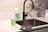 A modern kitchen sink setup with a sleek, black faucet. Next to the sink, there are two cleaning product bottles with a light green liquid inside. A gray cleaning cloth is draped on the counter, and there are a couple of small puddles of liquid near the bottles. In the background, a vase with eucalyptus branches adds a touch of greenery.