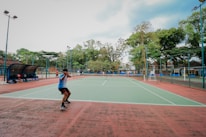 A person playing tennis on an outdoor court surrounded by tall trees and some seating areas covered with canopies. The sky appears partly cloudy, providing a calm backdrop for the action. The court has a green surface with red borders.