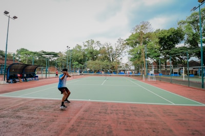 A person playing tennis on an outdoor court surrounded by tall trees and some seating areas covered with canopies. The sky appears partly cloudy, providing a calm backdrop for the action. The court has a green surface with red borders.