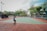 A person playing tennis on an outdoor court surrounded by tall trees and some seating areas covered with canopies. The sky appears partly cloudy, providing a calm backdrop for the action. The court has a green surface with red borders.