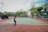 A person playing tennis on an outdoor court surrounded by tall trees and some seating areas covered with canopies. The sky appears partly cloudy, providing a calm backdrop for the action. The court has a green surface with red borders.