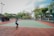A person playing tennis on an outdoor court surrounded by tall trees and some seating areas covered with canopies. The sky appears partly cloudy, providing a calm backdrop for the action. The court has a green surface with red borders.