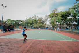 A person playing tennis on an outdoor court surrounded by tall trees and some seating areas covered with canopies. The sky appears partly cloudy, providing a calm backdrop for the action. The court has a green surface with red borders.