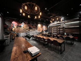 A spacious room with long wooden tables and chairs arranged for a meeting or conference. The walls are adorned with a large, colorful mural featuring intricate designs and a prominent portrait of a person's face. The lighting is dim, with several warm-toned lights hanging from circular chandeliers resembling wagon wheels. A white projector is placed on one of the tables, ready for use.