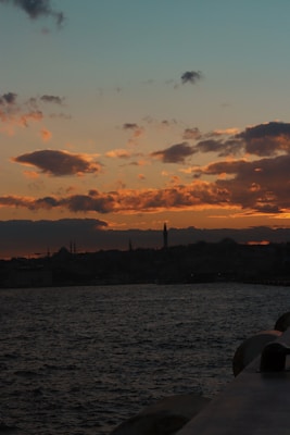 A scenic view of the horizon during sunset, with dark silhouettes of buildings and a minaret against a vibrant, colorful sky displaying hues of orange and pink. The foreground features a calm water body reflecting the warm colors of the sky.