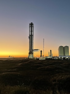 A towering rocket launch tower stands prominently against a twilight sky. The silhouette of a rocket is visible next to the tower, with additional industrial structures nearby. The horizon features a bright gradient from orange to blue, suggesting either dawn or dusk. The foreground consists of dark grass or vegetation.