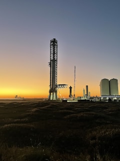 A towering rocket launch tower stands prominently against a twilight sky. The silhouette of a rocket is visible next to the tower, with additional industrial structures nearby. The horizon features a bright gradient from orange to blue, suggesting either dawn or dusk. The foreground consists of dark grass or vegetation.
