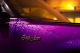 A close-up view of a car door with the emblem 'Royal Salon' prominently displayed. The car's surface is covered with water droplets, reflecting the surrounding lights. The paint is a vibrant purple, contrasting with warm yellow and orange ambient lighting, creating a striking visual effect.