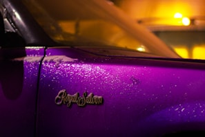 A close-up view of a car door with the emblem 'Royal Salon' prominently displayed. The car's surface is covered with water droplets, reflecting the surrounding lights. The paint is a vibrant purple, contrasting with warm yellow and orange ambient lighting, creating a striking visual effect.