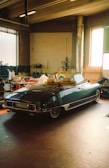 A classic convertible car is parked inside a spacious workshop. The vehicle has a sleek design with a dark green color. There are cardboard boxes and various tools scattered around, suggesting a space used for automotive repair or restoration. Large windows allow natural light to enter, illuminating the tiled floor and giving a warm ambiance.