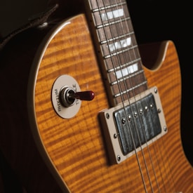 A close-up of an electric guitar's body featuring a wooden finish with a sunburst pattern, highlighting the grain. The image shows a pickup and a switch labeled 'Rhythm' and 'Treble'. Metal strings are visible, running along the neck towards the fretboard.