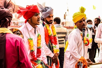 A group of men dressed in colorful traditional attire are engaged in a lively musical performance. They wear turbans in shades of red, yellow, and grey, adorned with vibrant garlands. One man is energetically singing into a microphone while playing a harmonium, surrounded by others who appear to be singing or playing music.