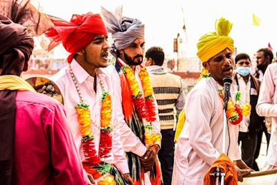 A group of men dressed in colorful traditional attire are engaged in a lively musical performance. They wear turbans in shades of red, yellow, and grey, adorned with vibrant garlands. One man is energetically singing into a microphone while playing a harmonium, surrounded by others who appear to be singing or playing music.