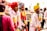A group of men dressed in colorful traditional attire are engaged in a lively musical performance. They wear turbans in shades of red, yellow, and grey, adorned with vibrant garlands. One man is energetically singing into a microphone while playing a harmonium, surrounded by others who appear to be singing or playing music.