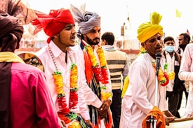 A group of men dressed in colorful traditional attire are engaged in a lively musical performance. They wear turbans in shades of red, yellow, and grey, adorned with vibrant garlands. One man is energetically singing into a microphone while playing a harmonium, surrounded by others who appear to be singing or playing music.