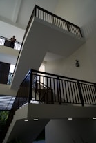An interior view of a building showcasing multiple levels of staircases with sleek, dark railings against white walls. Natural light filters in from above, highlighting the minimalistic and modern architectural design. A person is visible on one of the upper levels, leaning on the railing.