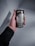 A hand is holding a can of Starbucks Doubleshot Espresso. The can is silver and black with the Starbucks logo and text in both English and Arabic. The person holding the can is wearing a black long-sleeve garment and a black beaded bracelet. The background is plain and gray.