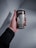 A hand is holding a can of Starbucks Doubleshot Espresso. The can is silver and black with the Starbucks logo and text in both English and Arabic. The person holding the can is wearing a black long-sleeve garment and a black beaded bracelet. The background is plain and gray.
