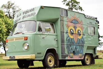 A vintage green camper van parked on grass, featuring a large, colorful owl mural painted on its side. The vehicle has a retro design, with round headlights and a roof extension displaying the words 'La Volva Sin Rumbo'. A scenic background of trees and sky enhances the tranquil setting.