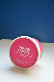 A round container with a pink lid featuring the brand name 'DOUGH LICIOUS' in bold white letters. Below the name, there are phrases like 'edible and bakeable cookie-dough' and '#lovelocal'. It rests on a white surface with a blue background.