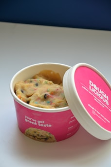 An open container of cookie dough with colorful specks sits on a white surface. The container is pink and features the brand name 'Dough Licious' along with the phrase 'you've got great taste.' The lid is resting partially open beside the container.