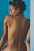 A woman with short, curly hair is sitting with her back facing the camera against a deep blue background. Her skin is smooth and slightly tanned, and her posture is relaxed.
