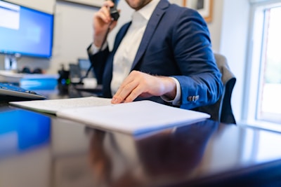A person in a suit is seated at a desk, holding a phone to their ear and using the other hand to turn pages in a document. The workspace includes a computer monitor, paperwork, and other office supplies. Large windows allow natural light to illuminate the area.