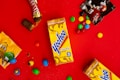A bright red background featuring a Yoo-hoo chocolate drink box, partially unwrapped Twix candy, assorted M&M's candies scattered around, and opened M&M's wrappers. The arrangement is playful and colorful, giving a sense of indulgence and fun.