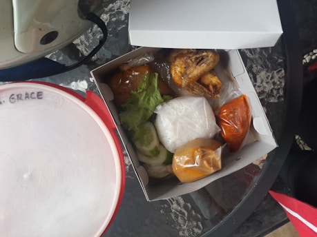 A takeout box containing various food items including fried chicken, sauce packets, and vegetables like lettuce and cucumber. The box is placed on a glass table along with another container labeled with 'GRACE'. The setting appears casual and serves as a meal setup.
