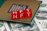 Red blocks with the letters NFT written on them are placed on a computer chip, overlaid on US dollar bills.