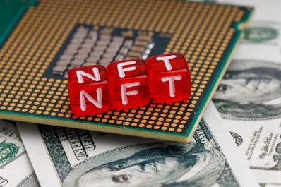 Red blocks with the letters NFT written on them are placed on a computer chip, overlaid on US dollar bills.