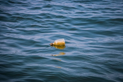 A plastic bottle floats on the surface of a body of water, with gentle ripples surrounding it. The water appears to be clean and blue.