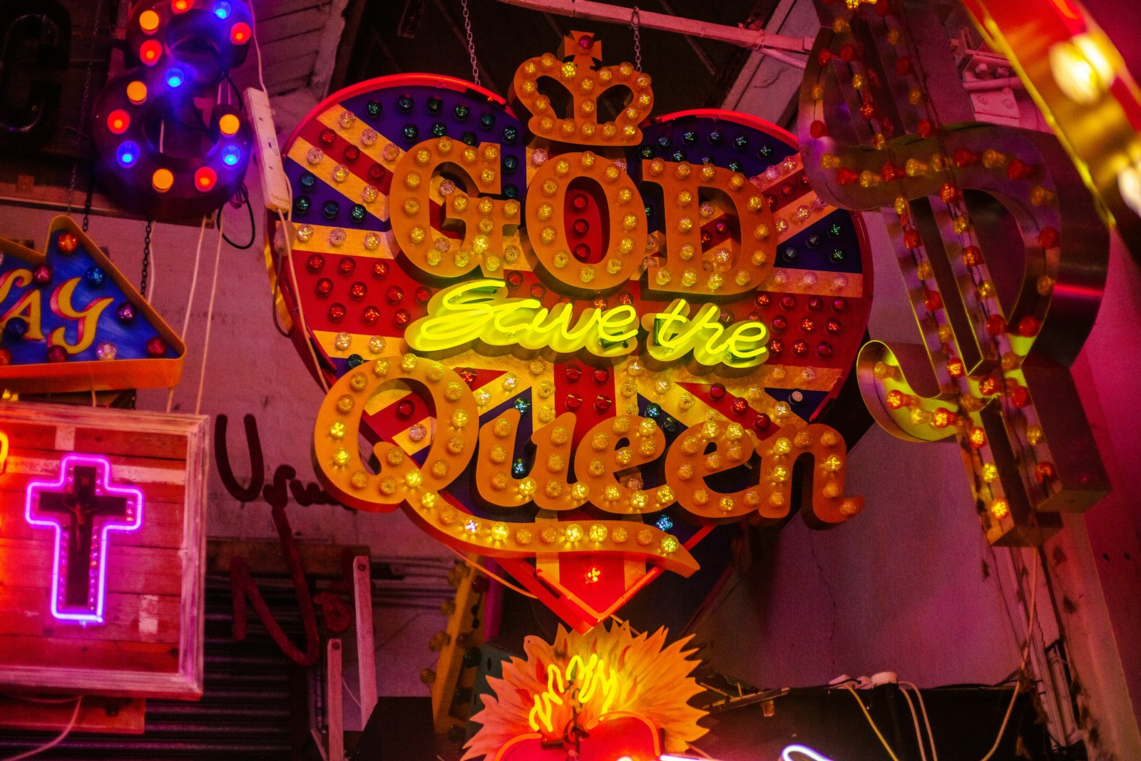 A heart-shaped Union Jack sign reading God Save the Queen in fairground bulbs, surrounded by glowing neon crosses and lettering on a warehouse wall
