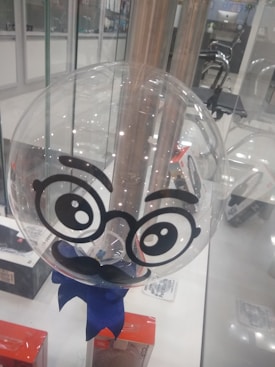 A transparent balloon with cartoonish eyes, eyebrows, and a mustache drawn on it. It sits on a display counter surrounded by various boxed items and is tied with a blue ribbon at the bottom. The background features a glass storefront with metal fixtures and office equipment visible.