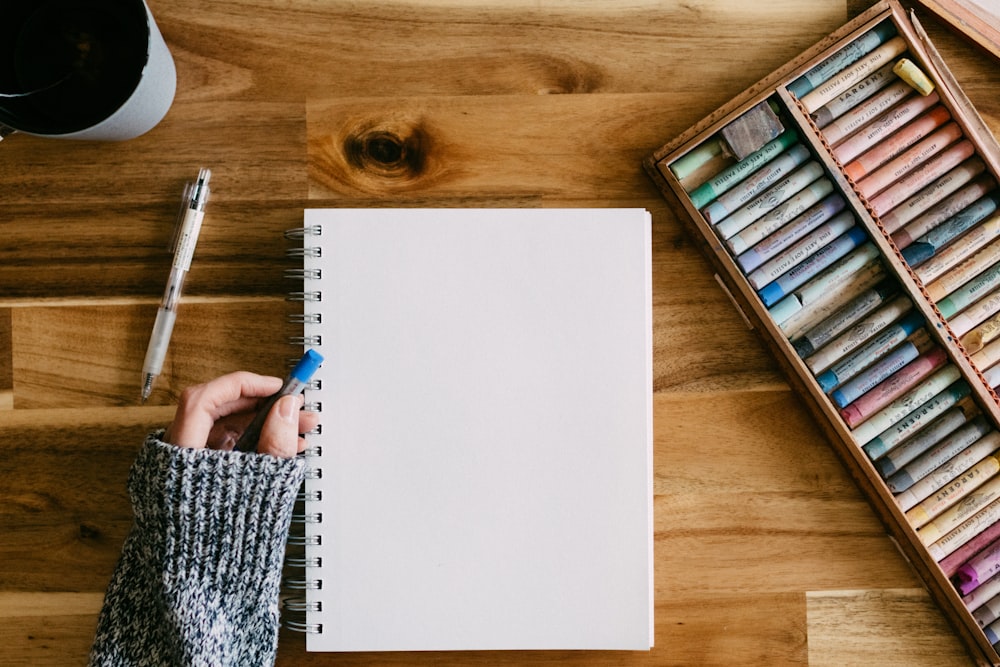 a person writing on a notebook surrounded by crayons