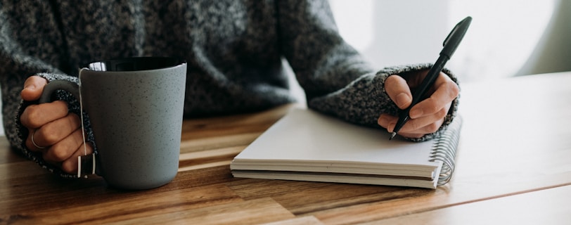 a person holding a pen and writing on a notebook with a mug of tea in the other hand