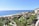 A resort-like coastal area features modern buildings with terracotta roofs nestled among lush green palm trees and landscaping. The scene overlooks a sandy beach meeting the calm, vast ocean under a clear blue sky.