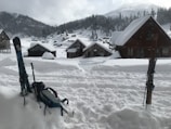 A snowy village landscape with wooden cabins partially covered in snow. Skis and a backpack are stuck upright in the deep snow, suggesting a winter sports activity. Pine trees are scattered across the hills, and a mountain range is visible in the distance under a cloudy sky.