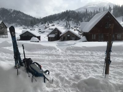 A snowy village landscape with wooden cabins partially covered in snow. Skis and a backpack are stuck upright in the deep snow, suggesting a winter sports activity. Pine trees are scattered across the hills, and a mountain range is visible in the distance under a cloudy sky.