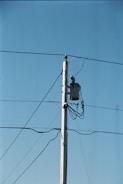 A wooden utility pole stands tall against a clear blue sky, with several wires extending from it. A transformer is mounted near the top of the pole, and multiple cables are wrapped around it.