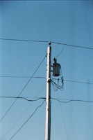 A wooden utility pole stands tall against a clear blue sky, with several wires extending from it. A transformer is mounted near the top of the pole, and multiple cables are wrapped around it.